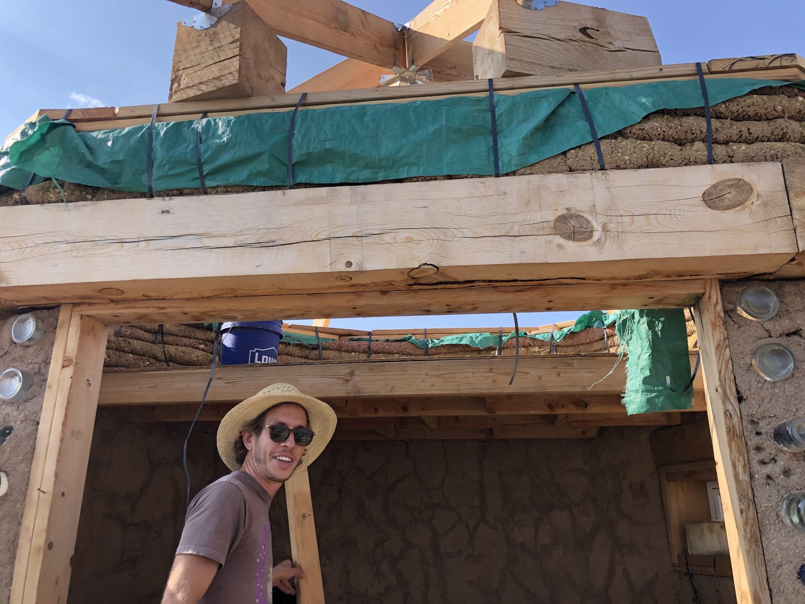 John smiling beneath a heavy timber lintel, with glass bottle bricks visible in the earthen walls on either side
