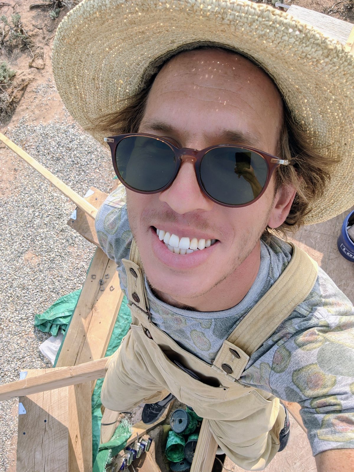 Selfie from atop wooden scaffolding, wearing work gloves, a straw hat, and overalls